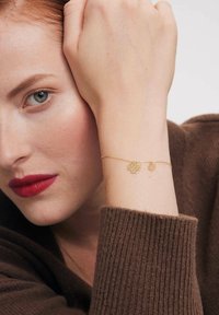 Złota bransoletka z florystycznym wzorem, z dwoma zawieszkami. Noszona na nadgarstku o jasnej karnacji, w zestawieniu z brązowym, dzianinowym swetrem.