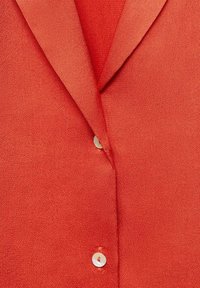 Gros plan sur un tissu rouge texturé avec un col, deux boutons blancs et un décolleté en V.