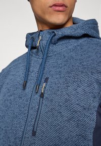 Μπλε φλις φούτερ με υφή, με φερμουάρ μπροστά, ρυθμιζόμενο σκουφάκι με κορδόνια και μαύρες λεπτομέρειες στα μανίκια.