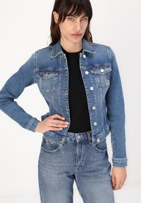 Giacca in denim blu medio, con due tasche sul petto e chiusure a bottoni argentati, abbinata a jeans in denim a vita alta con accenti di glitter.