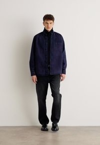 Cămașă din denim bleumarin peste un pulover cu guler înalt negru, asortată cu blugi închiși și pantofi negri. Cămașa dispune de un buzunar pe piept și închidere cu nasturi.