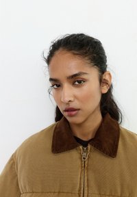 Ung kvinde med mørkt hår trukket tilbage, iført en tan jakke med en brun fløjlsagtig krave, der kigger direkte ind i kameraet mod en ensfarvet hvid baggrund.