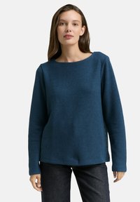 Blaues Rundhalsoberteil mit strukturiertem Stoff, langen Ärmeln und Knopfakzenten an der Seite, kombiniert mit dunklen Jeans.