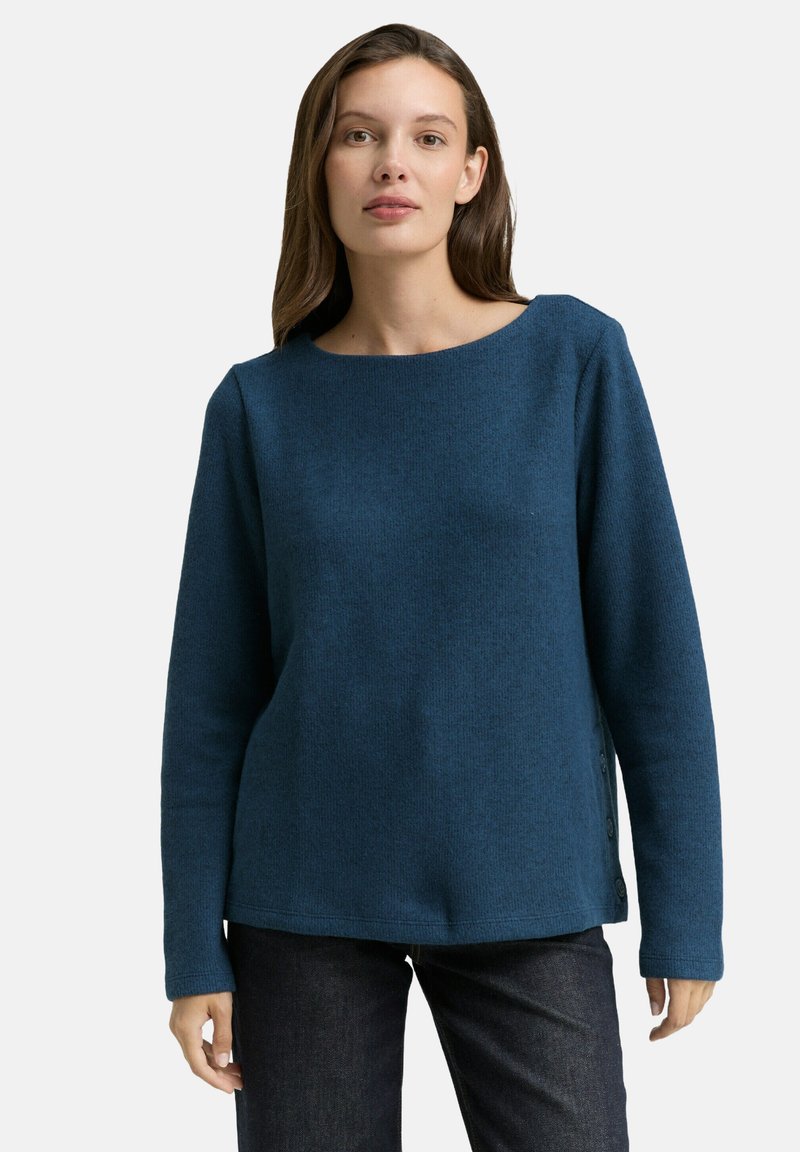 Blaues Rundhalsoberteil mit strukturiertem Stoff, langen Ärmeln und Knopfakzenten an der Seite, kombiniert mit dunklen Jeans.