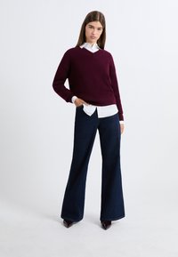 Bordowy sweter robiony na drutach z dekoltem w serek, noszony na białej koszuli z kołnierzykiem, w zestawieniu z dżinsami w ciemnym kolorze o rozszerzających się nogawkach i szpilkami o ostrym nosku.