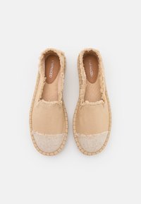 Une paire d'espadrilles en toile beige à enfiler avec des bords effilochés et une semelle tissée, vues de dessus sur un fond blanc.