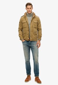 Bruine canvas jas met meerdere zakken en een capuchon, gedragen over een grijze coltrui. Lichtblauwe jeans en tan laarzen maken de outfit compleet.