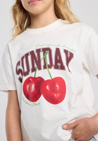 Camiseta de algodón blanca con texto en negrita morado que dice "DOMINGO" y un gráfico de dos cerezas rojas con tallos verdes. El texto dice "Cereza de California."