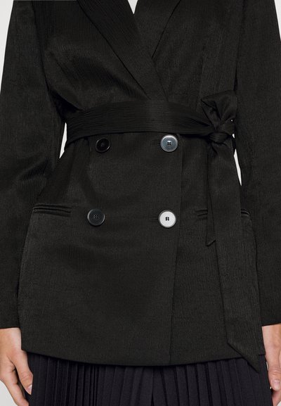 Zwarte textuur blazer met een strikceintuur, dubbel bevestigd ontwerp, drie ronde knopen (twee zwart, één wit) en smalle revers. Gepleat stof onderaan.