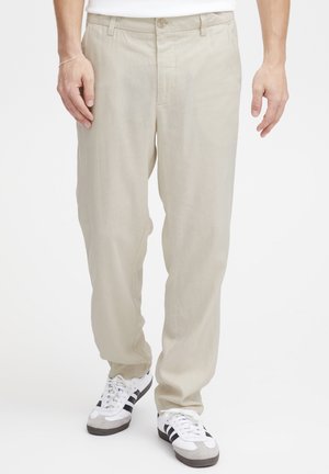 Homme portant un pantalon beige à coupe décontractée et des baskets blanches à rayures noires, debout devant un fond blanc uni.