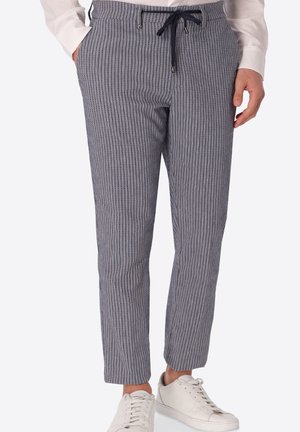 Uomo con pantaloni a righe blu e bianche con coulisse in vita, camicia bianca a maniche lunghe e sneakers bianche, mani in tasca.