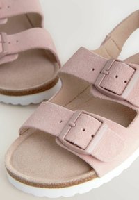 Roze suède sandaaltjes met twee verstelbare banden met bijpassende gespen, een gestructureerde voetbed en een witte rubberen zool met een golfpatroon.