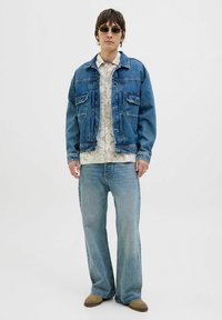 Chaqueta de mezclilla, azul claro, corte estándar con dos bolsillos en el pecho. Camisa blanca con estampado debajo, jeans azules y zapatos slip-on beige.