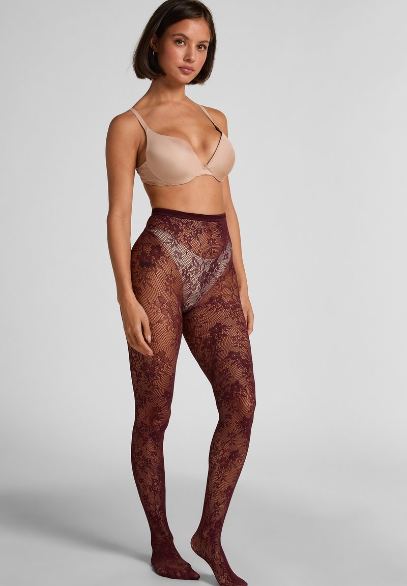 Collant in pizzo color bordeaux con motivo floreale e pannelli trasparenti, design a vita alta. Abbinati a un reggiseno color nude, la modella è in piedi davanti a uno sfondo neutro.