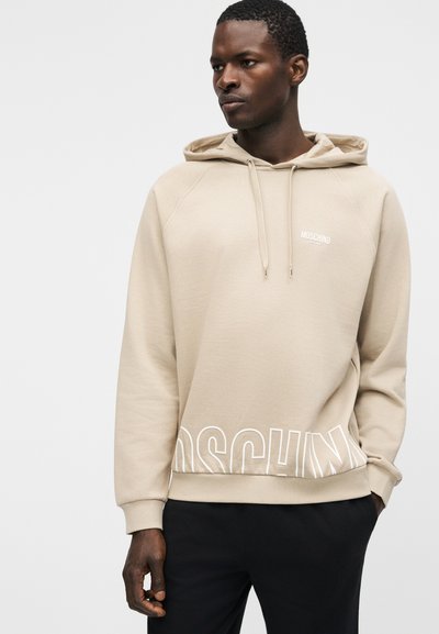 Homme portant un sweat à capuche beige Moschino avec cordons et logo partiel près de l'ourlet, associé à un pantalon noir, debout devant un fond uni.