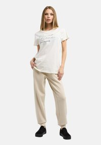 Weißes Baumwoll-T-Shirt mit Print und beigen Sweathosen. Das Model trägt schwarze Plateausneaker. Der Hintergrund ist schlicht grau.