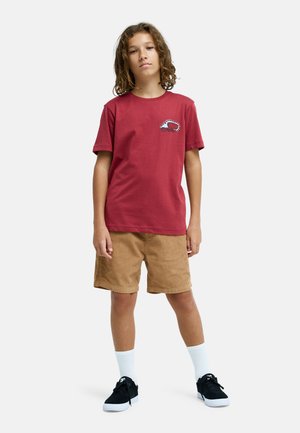 Jong persoon met krullend haar, gekleed in een rood T-shirt met print, beige korte broek, witte sokken en zwarte sneakers, staand tegen een witte achtergrond.