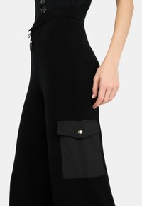 Weite schwarze Hosen aus weichem Stoff, mit einer großen Seitentasche und einem goldenen Druckknopf. Sanfte Textur, lässiges Design.