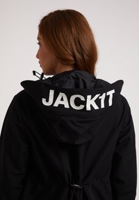 Svart vattentät jacka med huva, med en framträdande vit textlogotyp "JACK1T" på baksidan och justerbar dragsko i midjan.
