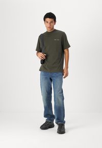 Tricou verde oliv cu mânecă scurtă, cu detalii subtile de text, purtat cu blugi albaștri estompați și teniși negri. Modelul poartă o geantă neagră de umăr.
