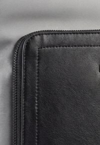 Estuche negro de cuero con cremallera y bordes cosidos sobre fondo de tela gris, mostrando primer plano de una esquina y detalles de la textura.