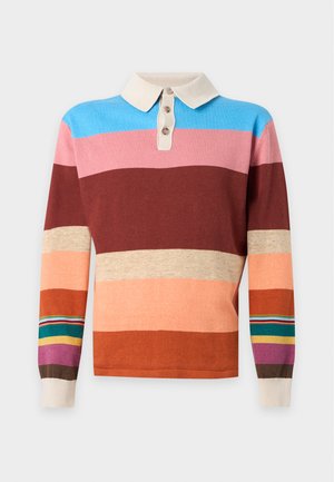 Homme debout portant un polo à rayures multicolores à manches longues, un pantalon noir et des baskets blanches sur un fond blanc uni.