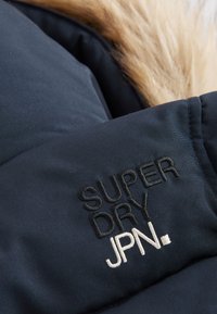 Tela de chaqueta navy con etiqueta bordada "SUPER DRY JPN." en blanco y negro. Presenta un cuello forrado de piel y textura acolchada.
