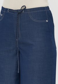 Blå denimbukser med snor i livet, frontlommer og kontrasterende hvit søm. Har en glatt tekstur og avslappet passform.