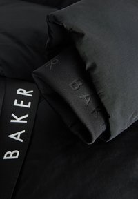 Gros plan sur la manche d'une veste matelassée noire avec un poignet côtelé montrant un texte embossé subtil et une sangle avec l'inscription blanche "BAKER".
