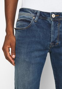 Tmavě modré džínové kalhoty s klasickým střihem. Mají páskové poutka, design s pěti kapsami a kontrastní hnědé prošití.