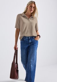 Beige poloshirt met een kraag, korte mouwen, gecombineerd met brede blauwe spijkerbroek. Bruine handtas met een hengsel in de hand. Soepele stoftextuur.