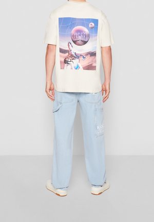 Beige T-Shirt mit einem bunten Grafiken auf dem Rücken, kombiniert mit hellblauen Cargo-Hosen und weißen Sneakern.