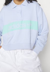 Ljust blå, croppad sweatshirt med polokrage. Har en mintgrön band med "adidas"-logotyp i en mjuk, matt finish.