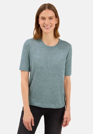 Lachende vrouw met schouderlang bruin haar, gekleed in een lichtblauwe, gestructureerde trui met korte mouwen en donkere broek, staande tegen een effen achtergrond.