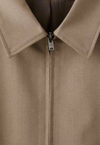 Giacca beige con colletto, dotata di una zip centrale con hardware metallico, realizzata in un tessuto testurizzato.