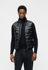 Άνδρας που φοράει μαύρο αμάνικο γιλέκο με καπιτονέ ύφασμα πάνω από μαύρο πουλόβερ με λαιμόκοψη και σκούρα τζιν, στέκεται μπροστά σε απλό λευκό φόντο.