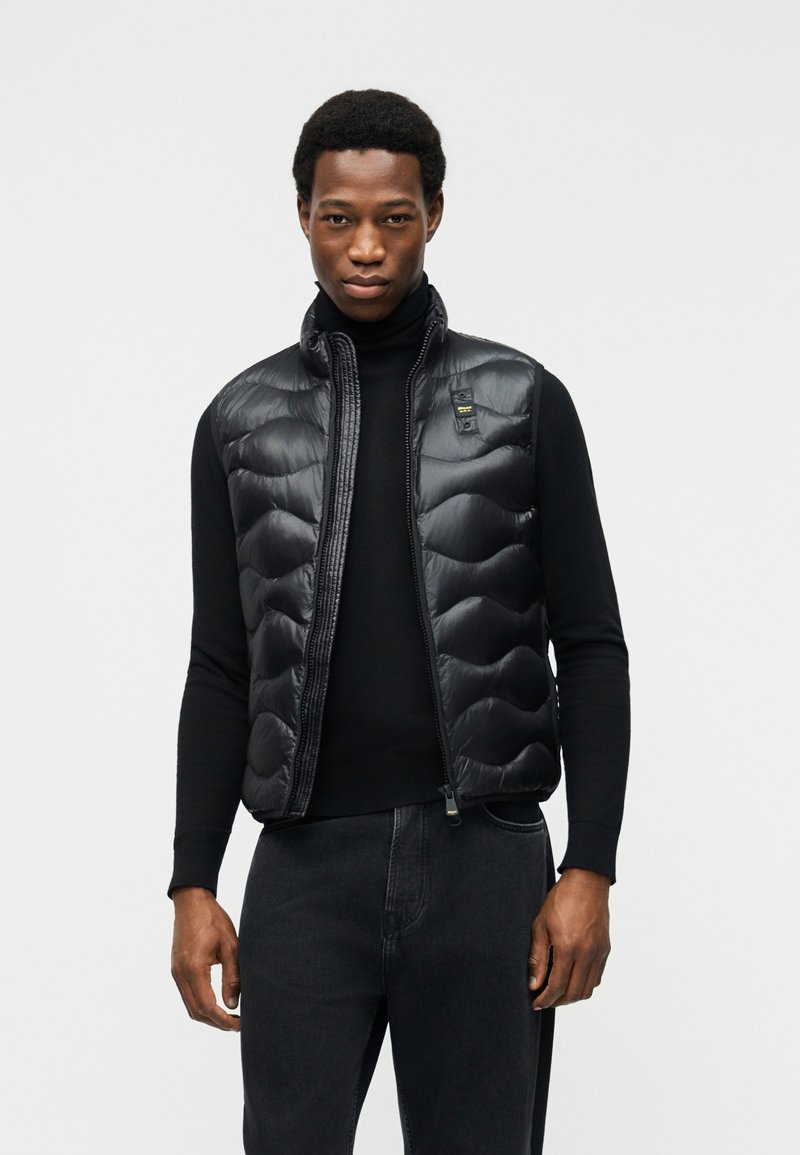 Άνδρας που φοράει μαύρο αμάνικο γιλέκο με καπιτονέ ύφασμα πάνω από μαύρο πουλόβερ με λαιμόκοψη και σκούρα τζιν, στέκεται μπροστά σε απλό λευκό φόντο.