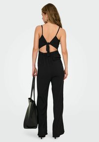 Zwarte jumpsuit met spaghettibandjes, een strik achteraan en een relaxte pasvorm met brede benen. Gecombineerd met een grote zwarte tote bag.