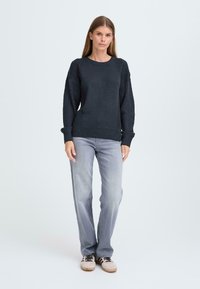 Donkergrijze gebreide trui met geribbelde manchetten en halslijn, gecombineerd met lichtgrijze rechtvallende jeans en beige sneakers. Glad textuur.