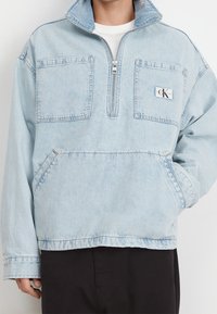 Ljusblå denimtröja med zippered hals, två framfickor och CK-logotyp. Har en avslappnad passform och synliga sömnadsdetaljer.