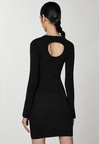 Abito nero a costine con maniche lunghe e scollatura tonda sulla schiena. Silhouette aderente che arriva fino a metà coscia. Tessuto liscio, design minimalista.