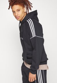 Felpa nera con cappuccio e accenti bianchi, con tre strisce sulle maniche, abbinata a pantaloni coordinati e una maglietta di un colore marrone chiaro sotto.