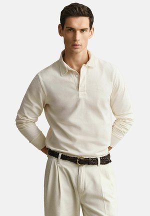 Junger Mann, der ein elfenbeinfarbenes Poloshirt mit langen Ärmeln trägt, in passende Hosen gesteckt, mit einem dunklen geflochtenen Gürtel, steht mit den Händen hinter dem Rücken.