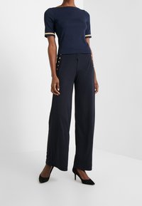 Tmavě modrý top s krátkými rukávy a zlatým lemováním, doplněný tmavě modrými širokými kalhotami se zlatými knoflíky po stranách, které jsou kombinovány s černými vysokými podpatky.