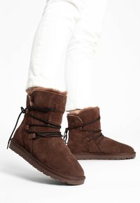 Stivaletti alla caviglia in suede marrone con foderato in pelliccia sintetica, dotati di lacci neri e suola in gomma. Design semplice e arrotondato con accenti minimi.