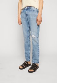 Ljusblå denimjeans med avslappnad passform och en trasig detalj vid knäet. Kombinerade med svarta sandaler med remmar.