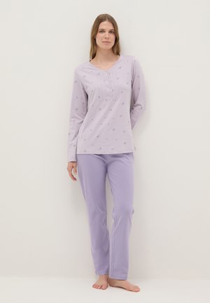 Donna che indossa una maglia a maniche lunghe viola chiaro con piccolo motivo floreale e pantaloni viola tinta unita abbinati, in piedi a piedi nudi contro una parete semplice.