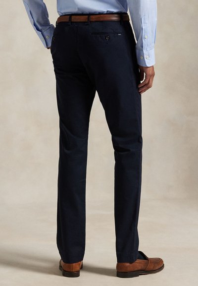 Muž oblečený v tmavě modrých kalhotách na míru, světle modré košili, hnědém pásku a hnědých kožených botách, stojící před neutrálním pozadím.