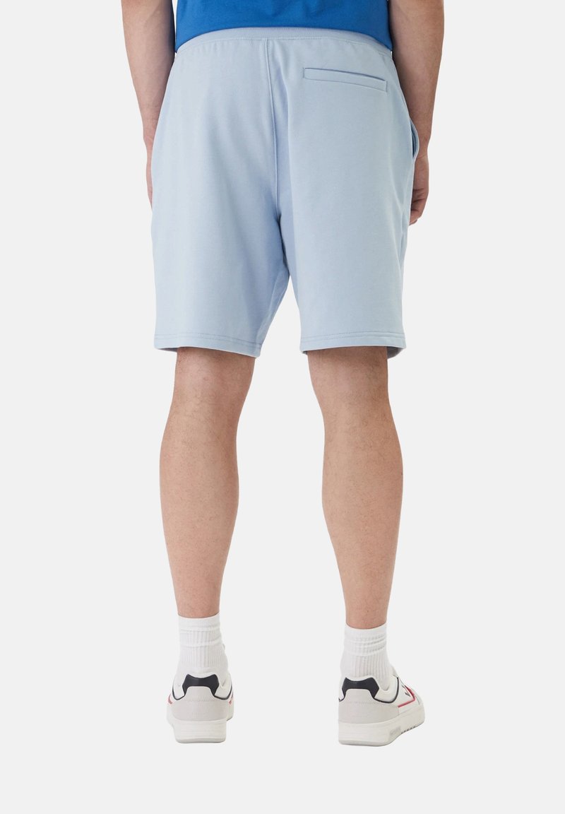 Homme portant un short bleu clair, des chaussettes blanches et des baskets blanches, debout de dos devant un fond uni.