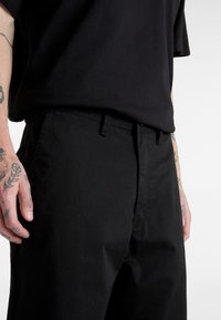 Schwarze Baumwollhosen mit geradem Schnitt, ausgestattet mit Seitentaschen und einem Reißverschluss. Nahaufnahme der Stoffstruktur und der Details des Bunds.